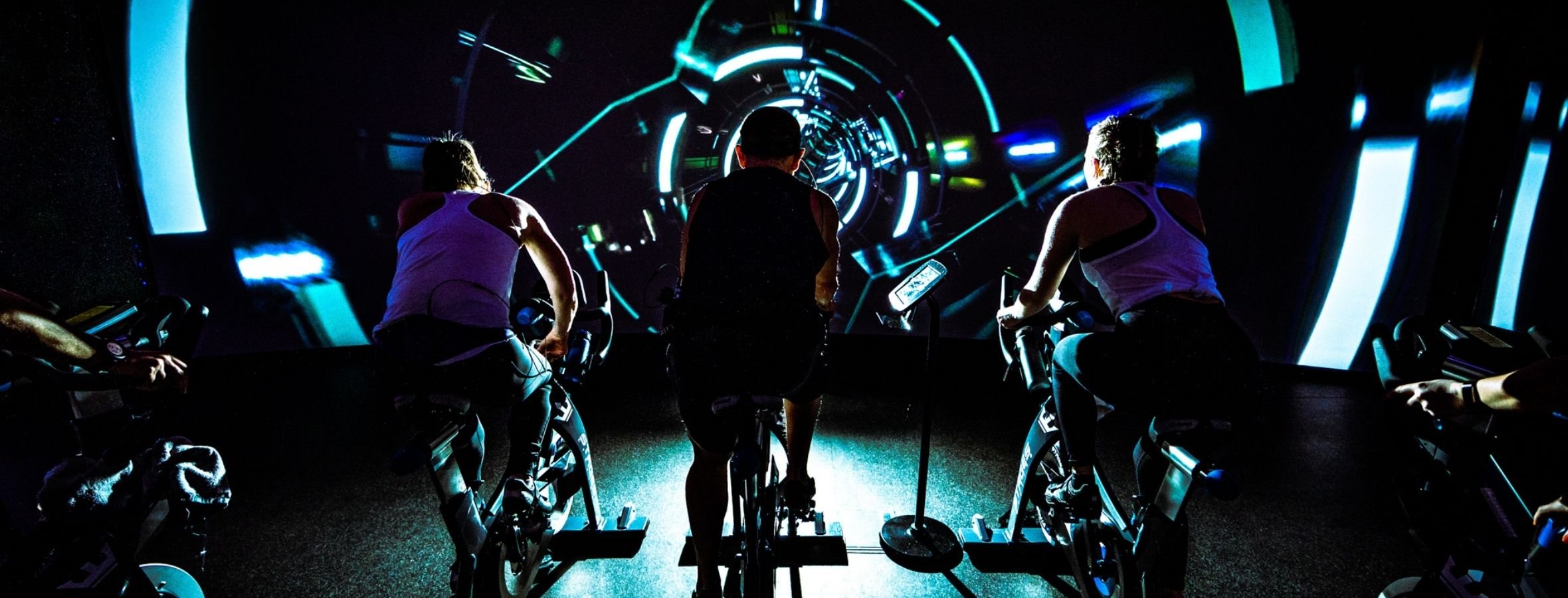 A virtual cycling studio with three cyclists and a dark futuristic scene on a video wall