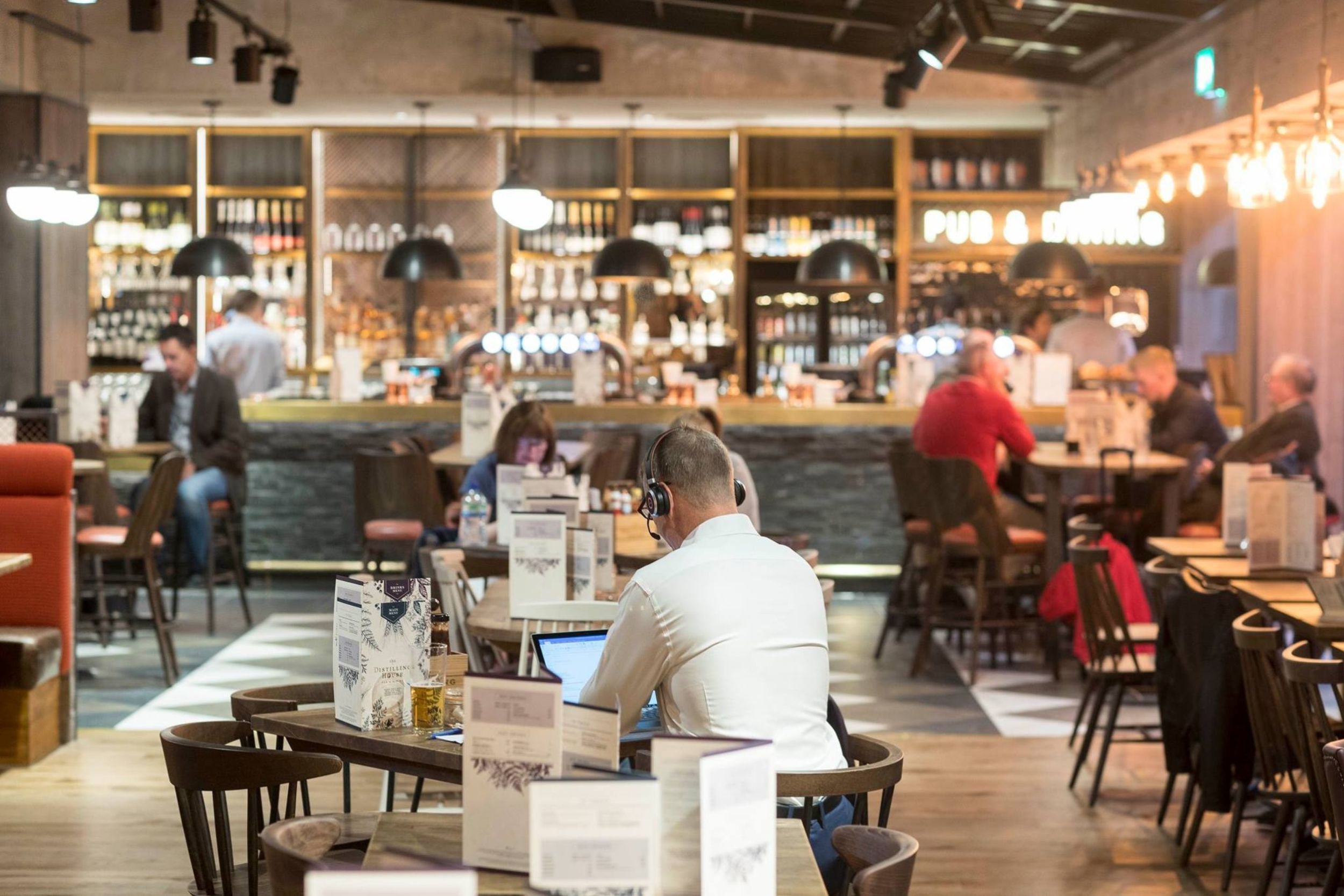 A restaurant with a discrete AV solution. People sitting at tables, some working on laptops, others relaxing. Bar in the background